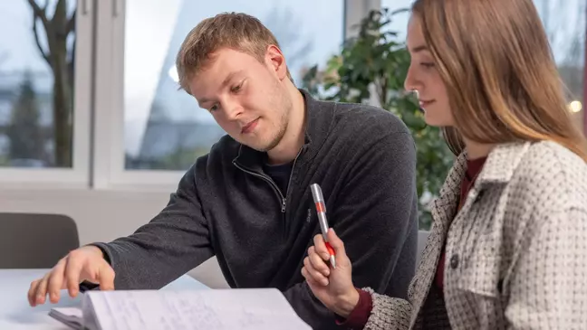 Ein Mann sitzt in einer modernen Räumlichkeit, er unterhält sich und hat ein Notizbuch geöffnet, er hält einen Stift