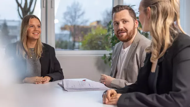 Dve mladé ženy a jeden mladý muž sedia pri stole a rozprávajú sa