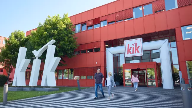 Červená budova KiK s veľkým bielym logom pred vchodom a niekoľko ľudí na ceste do budovy.