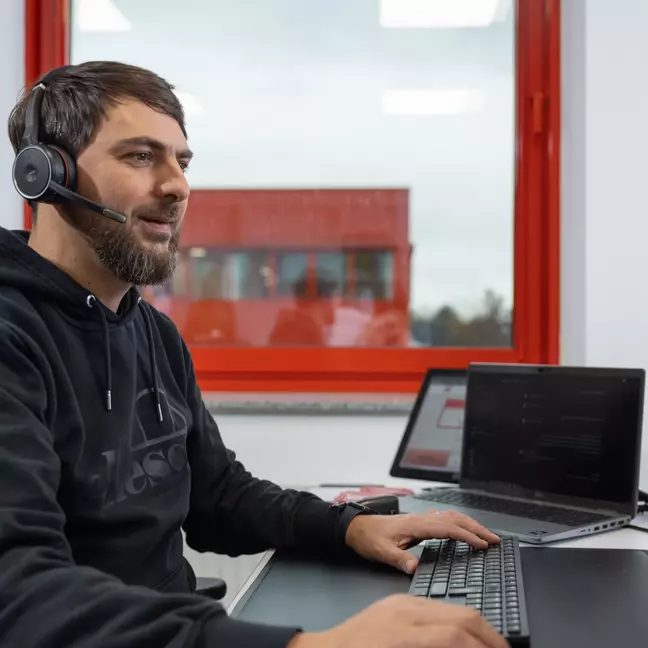 Ein Mann sitzt am Computer und hat ein Headset auf, im Hintergrund ist ein weiteres Gebäude