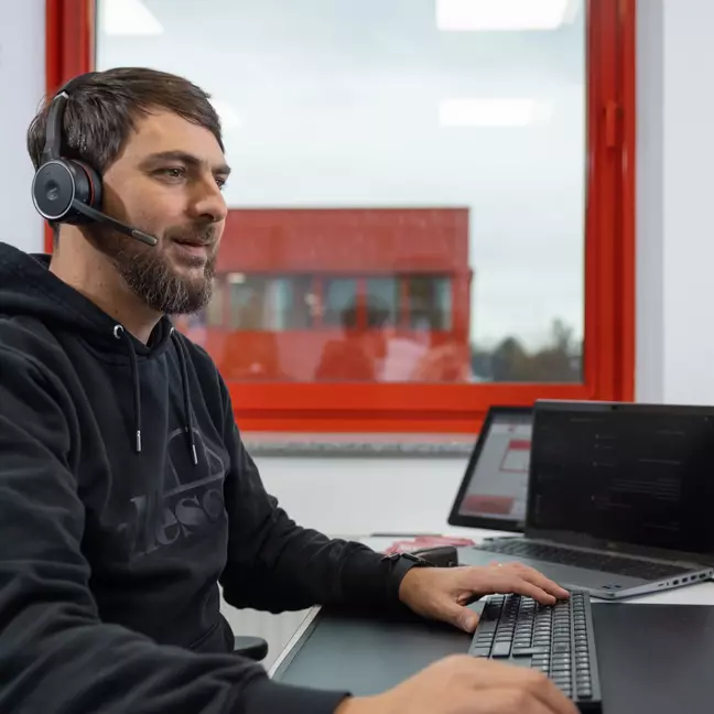 Ein Mann sitzt am Computer und hat ein Headset auf, im Hintergrund ist ein weiteres Gebäude